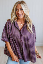 A smiling blonde woman wearing a loose purple and white plaid button-up shirt with light blue jeans, standing against a light neutral wall.