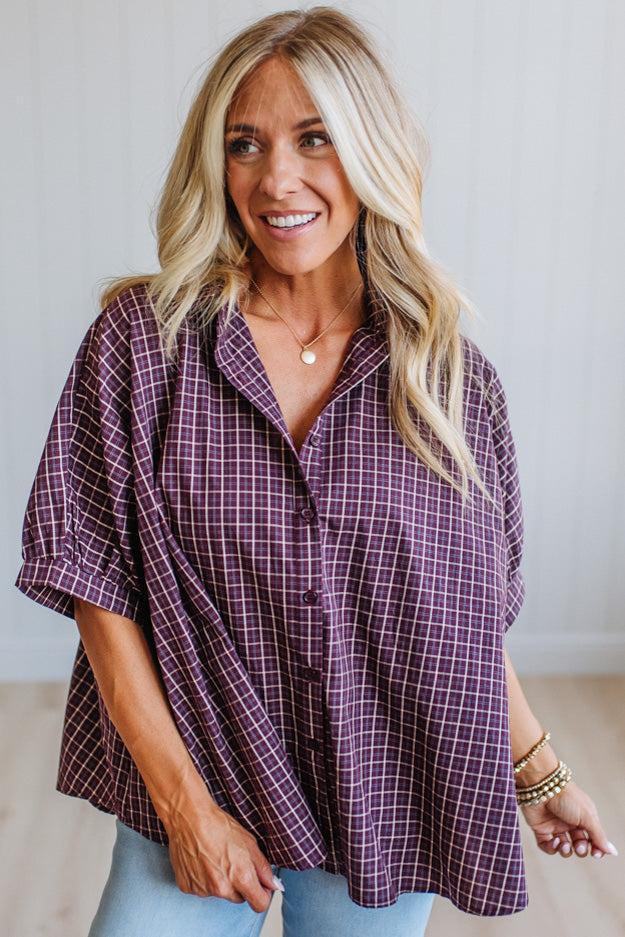 A smiling blonde woman wearing a loose purple and white plaid button-up shirt with light blue jeans, standing against a light neutral wall.