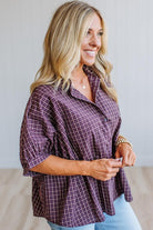 A woman wearing a purple and white plaid button-down shirt with ruffled sleeves, paired with light blue jeans, standing against a light gray wall.