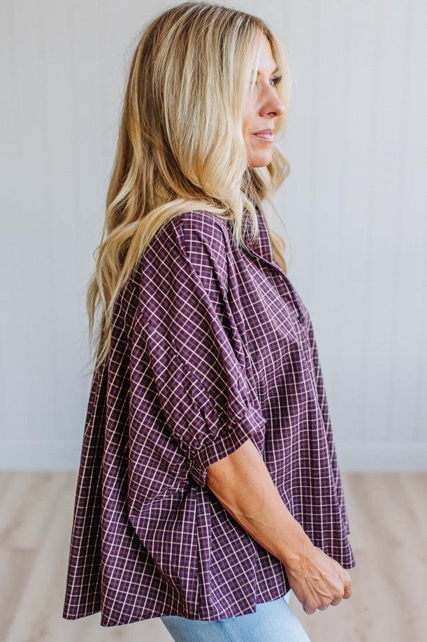 A woman with long blonde hair is shown in profile wearing a loose-fitting, short-sleeve button-up shirt with a purple and white plaid pattern, paired with light blue jeans, set against a neutral indoor background.