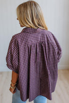 A woman shown from the back wearing a loose, short-sleeve button-up blouse with a purple and white windowpane plaid pattern, paired with light blue jeans.