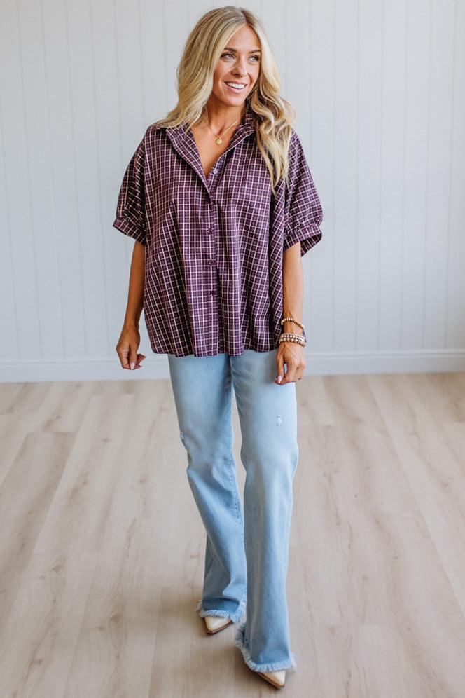 A woman modeling a short-sleeve button-up shirt with a purple and black plaid pattern, paired with light-wash blue jeans, standing in a room with light wood floors and white paneled walls.