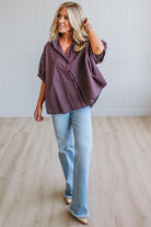 A woman modeling a short-sleeve button-up plaid shirt in dark purple and white, paired with light-wash blue jeans, standing on a light wood floor against a white paneled wall.