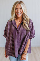 A smiling blonde woman wearing a loose purple and white plaid button-up shirt with light blue jeans, standing against a white paneled wall.