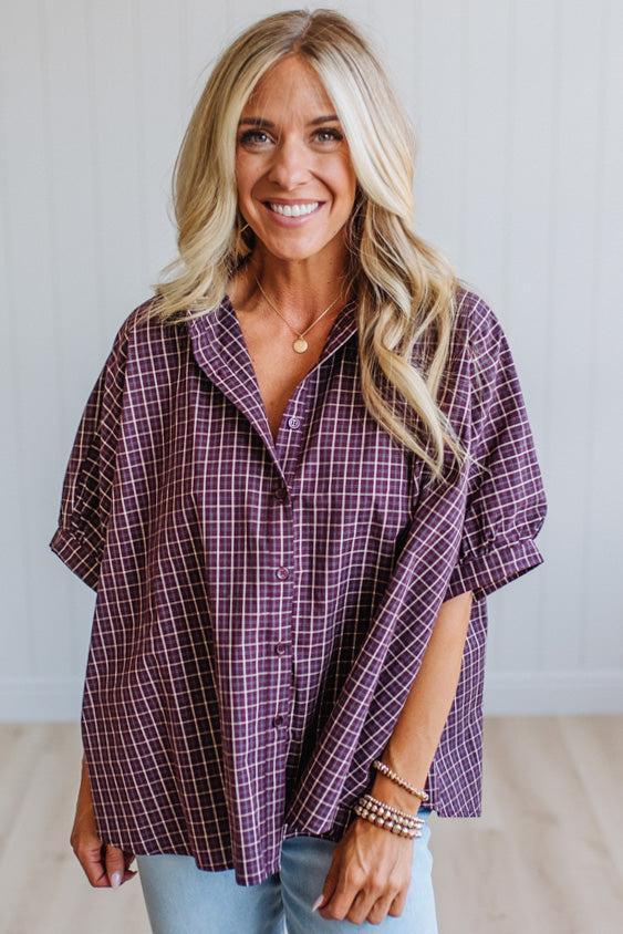 A smiling blonde woman wearing a loose purple and white plaid button-up shirt with light blue jeans, standing against a white paneled wall.