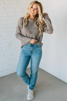 Woman wearing a gray sweater and blue jeans against a white brick wall.