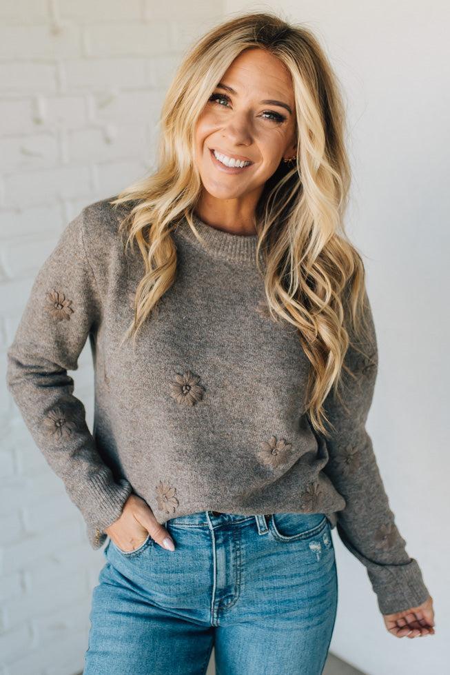 Woman wearing a brown sweater with floral details and blue jeans against a white brick wall.