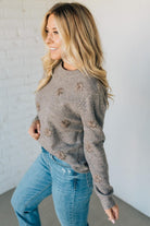 Woman wearing a gray sweater with floral patterns and blue jeans against a white wall.
