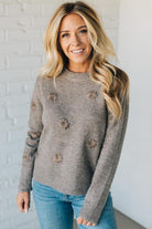 Woman wearing a gray sweater with floral patterns against a white brick wall.