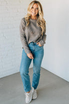 Woman wearing a gray sweater, blue jeans, and white sneakers against a white brick wall.