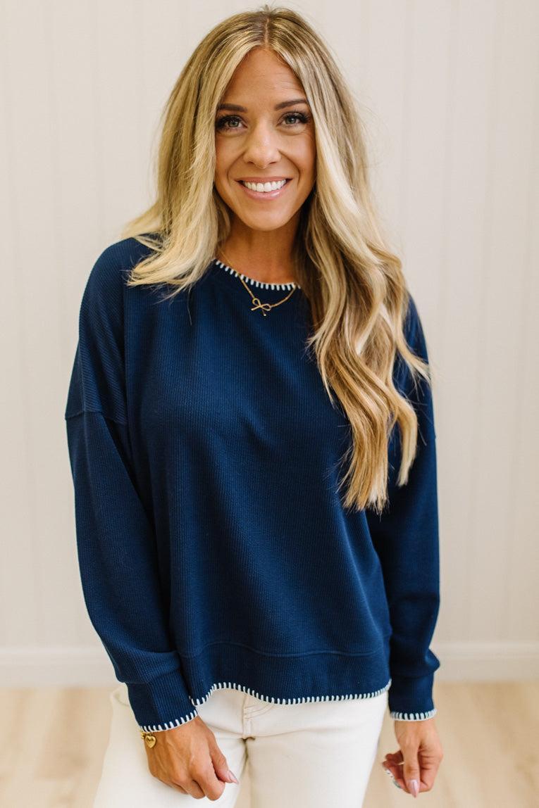 BLONDE WOMAN WEARING A WAFFLE TEXTURE TOP WITH A CREAM BLANKET STITCH TRIM, STANDING AGAINST A LIGHT WALL ON A LIGHT WOOD FLOOR. THE TOP IS NAVY.