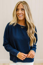 BLONDE WOMAN WEARING A WAFFLE TEXTURE TOP WITH A CREAM BLANKET STITCH TRIM, STANDING AGAINST A LIGHT WALL ON A LIGHT WOOD FLOOR. THE TOP IS NAVY.