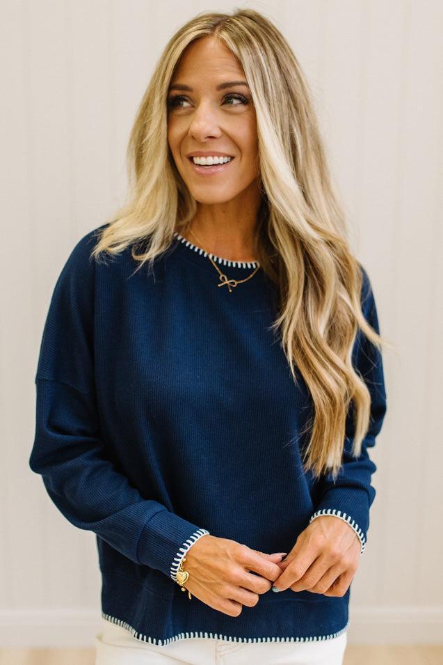BLONDE WOMAN WEARING A WAFFLE TEXTURE TOP WITH A CREAM BLANKET STITCH TRIM, STANDING AGAINST A LIGHT WALL ON A LIGHT WOOD FLOOR. THE TOP IS NAVY.