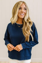 BLONDE WOMAN WEARING A WAFFLE TEXTURE TOP WITH A CREAM BLANKET STITCH TRIM, STANDING AGAINST A LIGHT WALL ON A LIGHT WOOD FLOOR. THE TOP IS NAVY.
