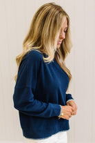BLONDE WOMAN WEARING A WAFFLE TEXTURE TOP WITH A CREAM BLANKET STITCH TRIM, STANDING AGAINST A LIGHT WALL ON A LIGHT WOOD FLOOR. THE TOP IS NAVY.