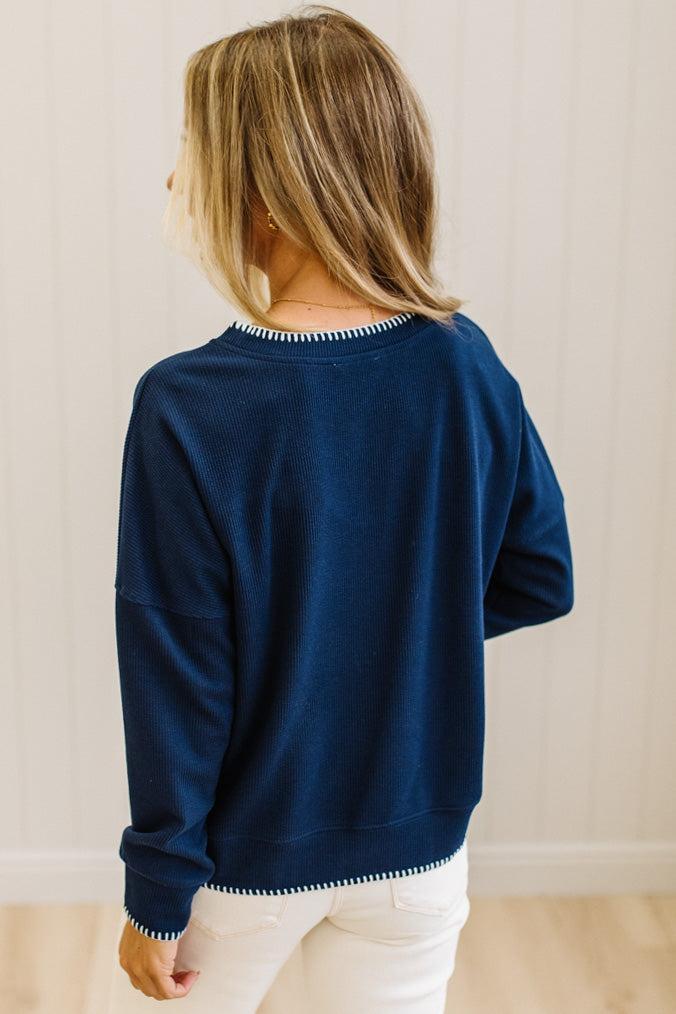 BLONDE WOMAN WEARING A WAFFLE TEXTURE TOP WITH A CREAM BLANKET STITCH TRIM, STANDING AGAINST A LIGHT WALL ON A LIGHT WOOD FLOOR. THE TOP IS NAVY.