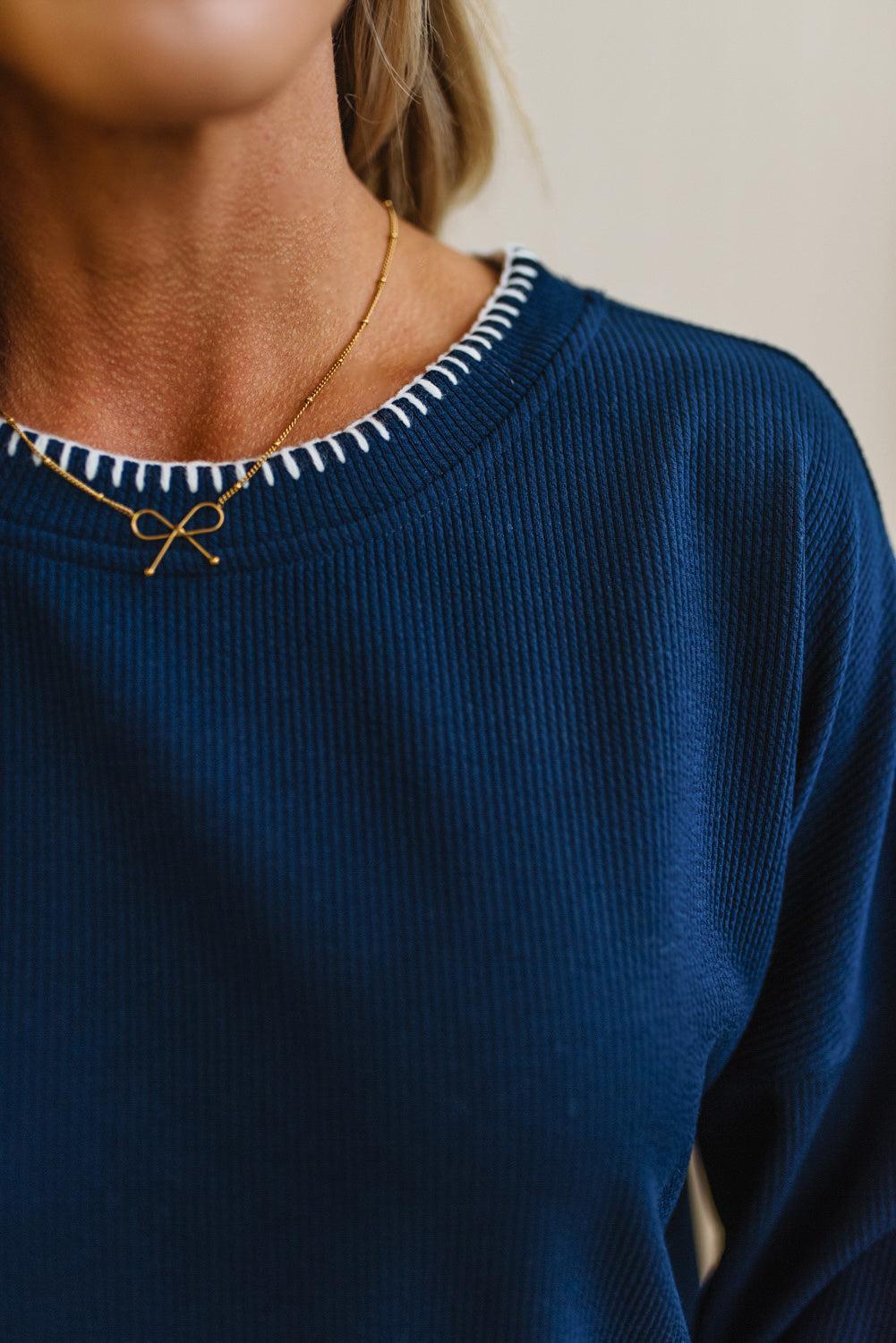 BLONDE WOMAN WEARING A WAFFLE TEXTURE TOP WITH A CREAM BLANKET STITCH TRIM, STANDING AGAINST A LIGHT WALL ON A LIGHT WOOD FLOOR. THE TOP IS NAVY.