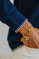 A close-up of a person's wrist wearing a gold chain bracelet with a heart charm, styled with a navy blue ribbed sweater and white pants.