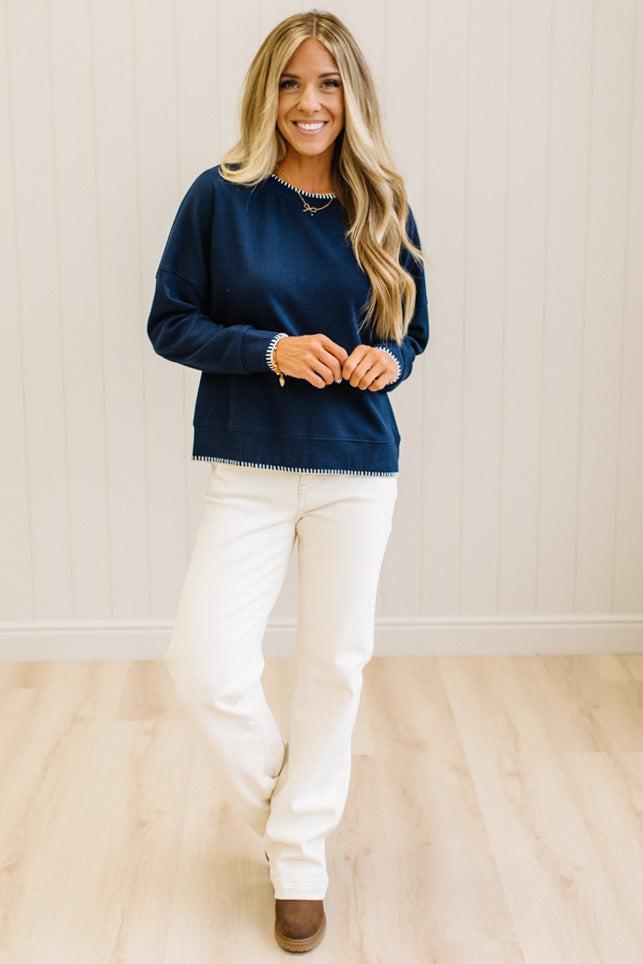 BLONDE WOMAN WEARING A WAFFLE TEXTURE TOP WITH A CREAM BLANKET STITCH TRIM, STANDING AGAINST A LIGHT WALL ON A LIGHT WOOD FLOOR. THE TOP IS NAVY.