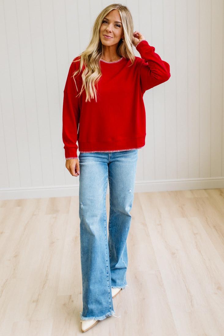 A woman modeling a bright red crewneck sweatshirt with white ruffle trim, paired with light-wash blue flare jeans and white pointed-toe flats, standing in a room with light wood floors and white paneled walls.