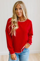 BLONDE WOMAN WEARING A WAFFLE TEXTURE TOP WITH A CREAM BLANKET STITCH TRIM, STANDING AGAINST A LIGHT WALL ON A LIGHT WOOD FLOOR. THE TOP IS RED.