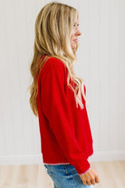 BLONDE WOMAN WEARING A WAFFLE TEXTURE TOP WITH A CREAM BLANKET STITCH TRIM, STANDING AGAINST A LIGHT WALL ON A LIGHT WOOD FLOOR. THE TOP IS RED.