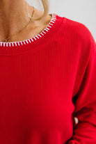 BLONDE WOMAN WEARING A WAFFLE TEXTURE TOP WITH A CREAM BLANKET STITCH TRIM, STANDING AGAINST A LIGHT WALL ON A LIGHT WOOD FLOOR. THE TOP IS RED.