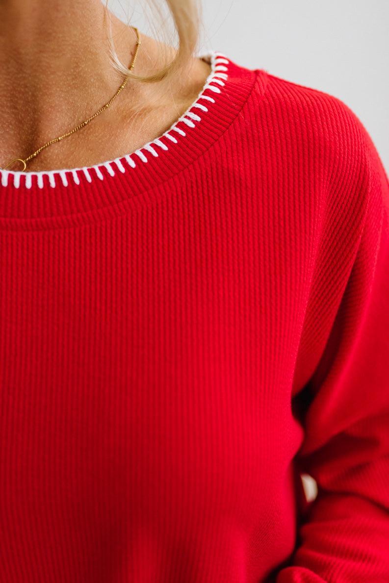 BLONDE WOMAN WEARING A WAFFLE TEXTURE TOP WITH A CREAM BLANKET STITCH TRIM, STANDING AGAINST A LIGHT WALL ON A LIGHT WOOD FLOOR. THE TOP IS RED.