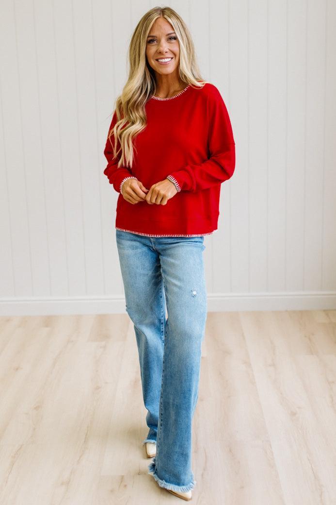 BLONDE WOMAN WEARING A WAFFLE TEXTURE TOP WITH A CREAM BLANKET STITCH TRIM, STANDING AGAINST A LIGHT WALL ON A LIGHT WOOD FLOOR. THE TOP IS RED.