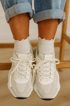White sneakers worn with light blue jeans and white socks on a wooden floor.
