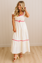Woman wearing a white dress with red trim against a light wooden paneled wall.