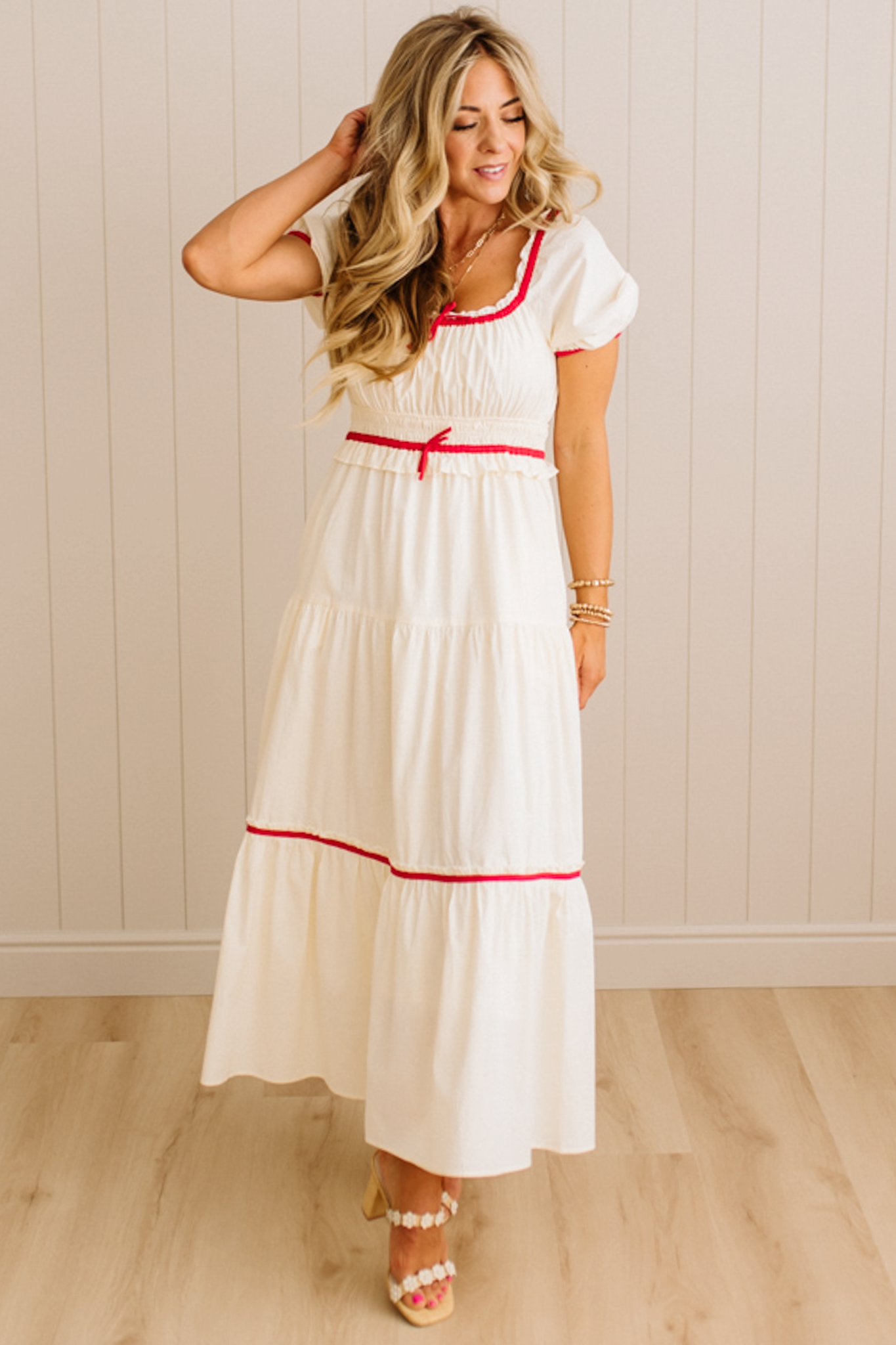 Woman wearing a white dress with red accents in a room with wooden flooring and a light-colored wall.