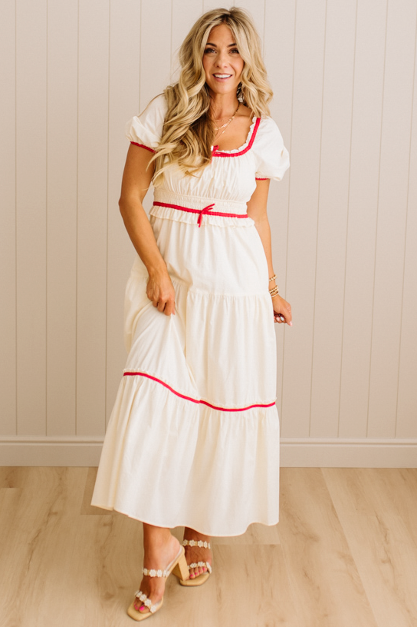 Woman wearing a white dress with red trim standing against a light wooden paneled wall.