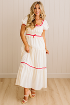 Woman wearing a white dress with red trim standing against a light wooden paneled wall.