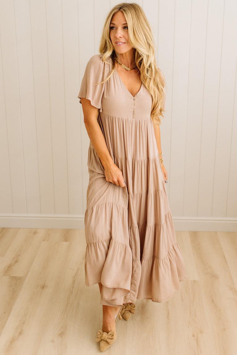 Woman wearing a long beige dress standing in a room with wooden flooring and white walls.
