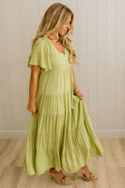 Woman wearing a light green tiered dress against a white paneled wall.
