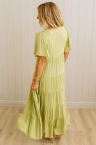 Woman wearing a light green dress with ruffled sleeves and bottom hem against a white paneled wall.