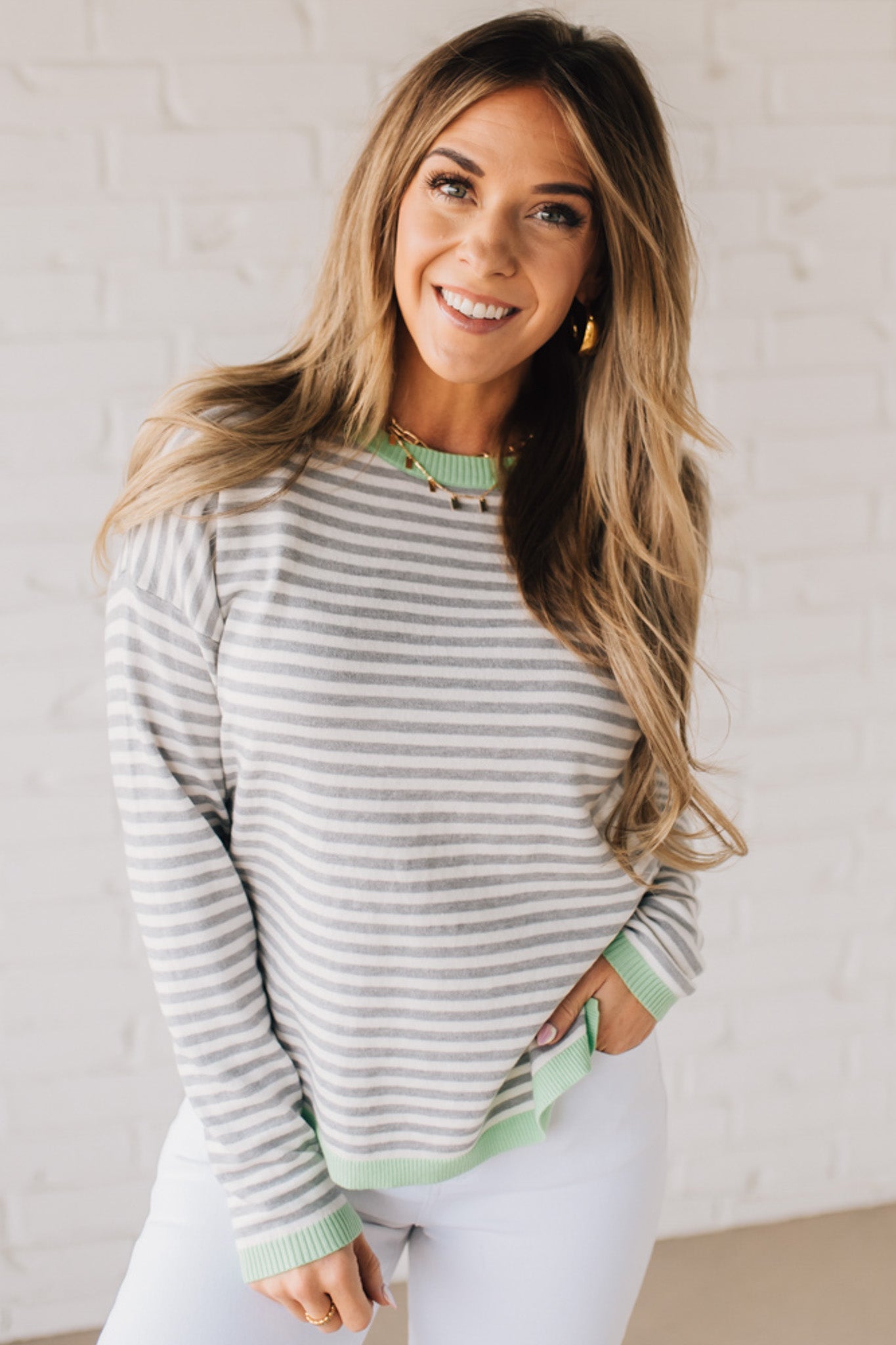 Blonde woman wearing a lightweight sweater with grey and white stripes with light green trim at neckline, wrist and hem.