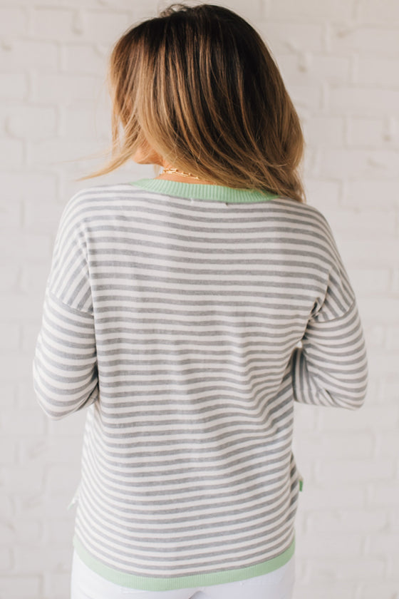 Blonde woman wearing a lightweight sweater with grey and white stripes with light green trim at neckline, wrist and hem.