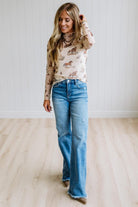 A woman wearing a beige long-sleeve turtleneck top with a western-themed horse print and blue flare jeans, standing in a room with light wood floors and white paneled walls.