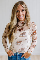 A smiling woman with long blonde hair wearing a beige long-sleeve mock neck top with a brown horse and cactus print, paired with blue jeans.