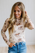 A woman wearing a beige long-sleeve mock-neck top with a western-themed horse and cow print, paired with blue jeans.