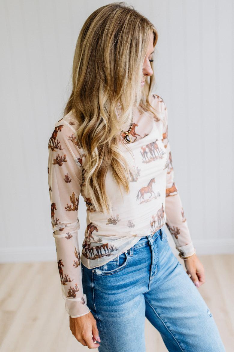 A woman wearing a beige long-sleeve t-shirt with a brown horse and cowboy print, paired with blue jeans, standing in a bright room.