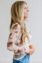 A woman wearing a beige long-sleeve top with a brown moose and deer print, paired with blue jeans, shown in a side-profile view against a white paneled wall.