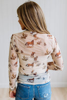 A woman viewed from behind wearing a beige long-sleeve top with a brown horse and cactus print, paired with blue jeans.