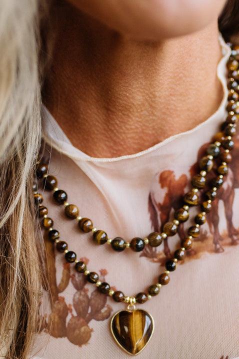 Close-up of a person wearing a double-strand beaded necklace with polished brown and black tiger's eye beads and a heart-shaped pendant with a gold-tone border and tiger's eye inlay.
