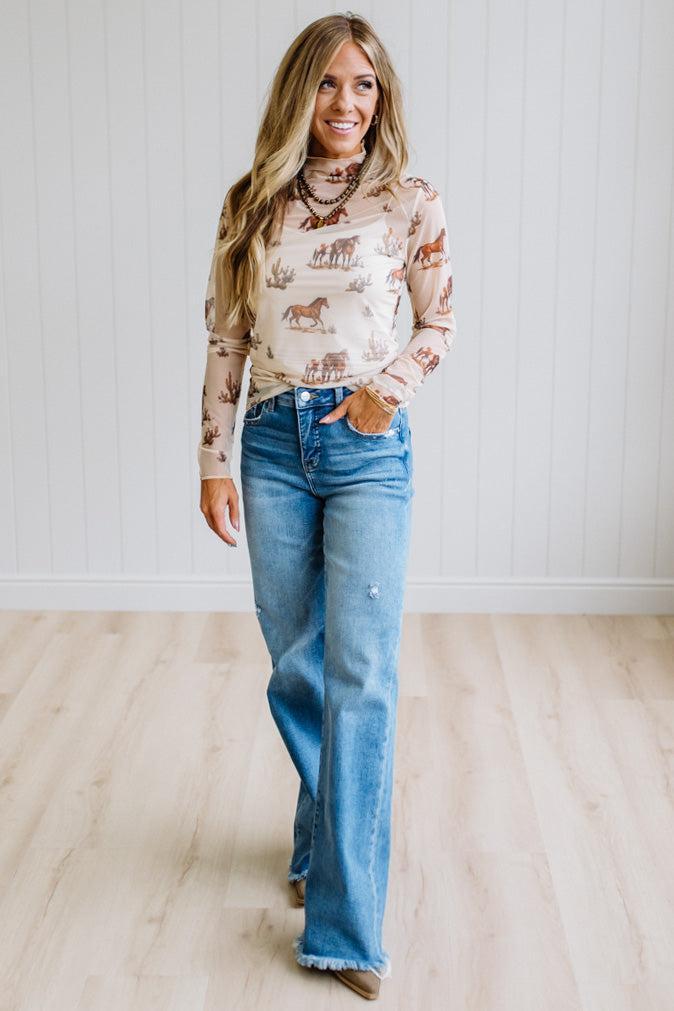 A woman modeling a beige long-sleeve mock-neck shirt with a brown horse and rider pattern, paired with blue jeans, standing in a room with light wood floors and white paneled walls.