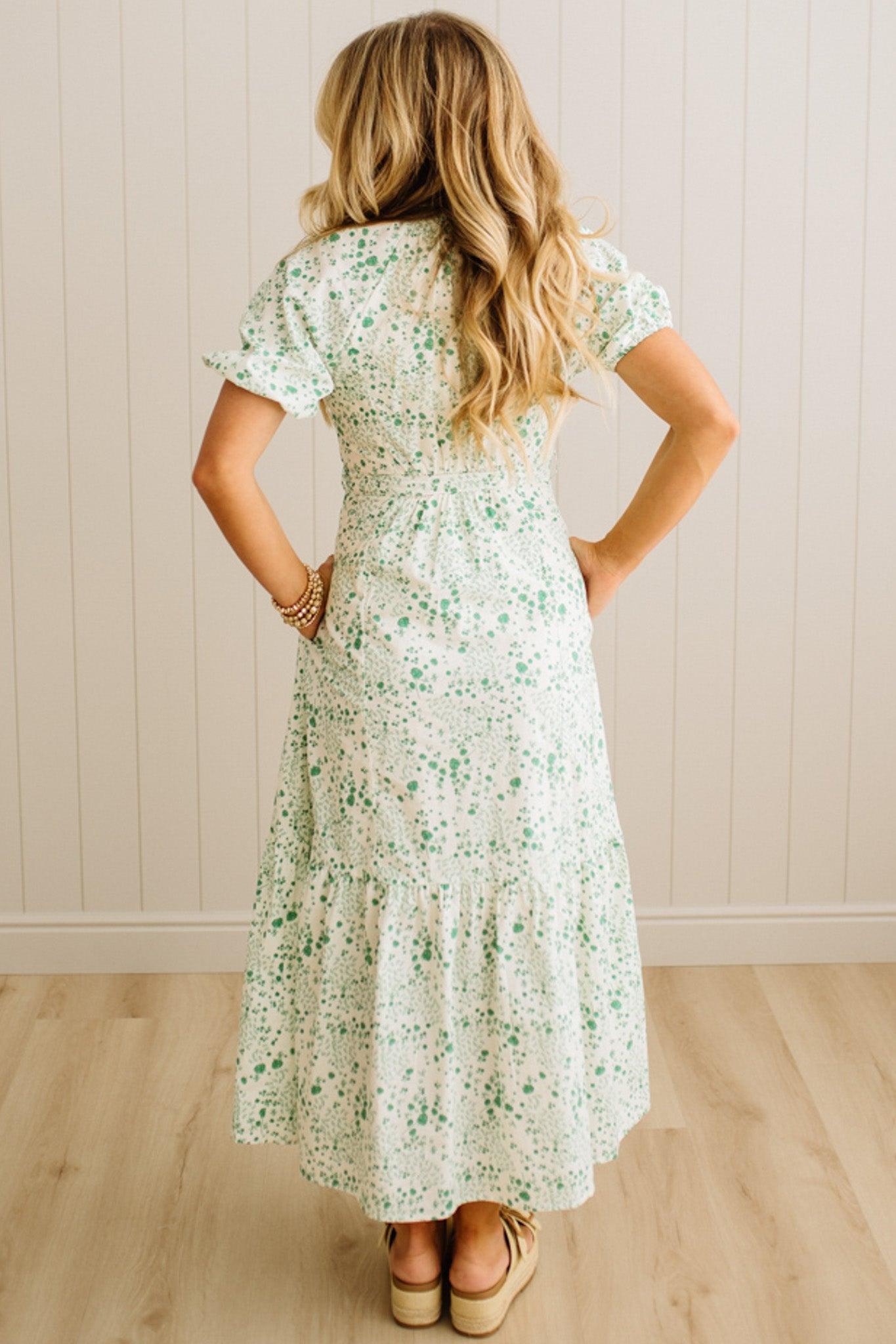 Woman wearing a floral dress standing against a light wooden paneled wall.