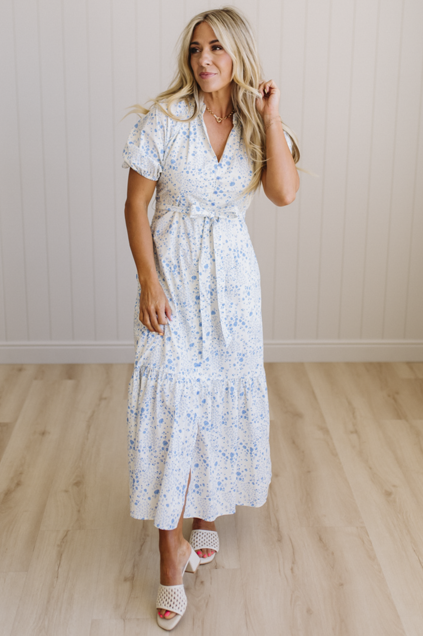 Woman wearing a floral dress standing in a room with wooden flooring and a light-colored wall.
