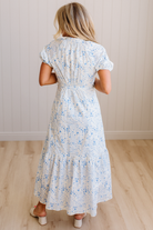 Woman wearing a white floral dress standing in a room with wooden flooring and a light-colored wall.